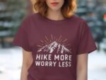 Woman wearing a maroon t-shirt with a mountain graphic and text "HIKE MORE WORRY LESS" against a natural background.