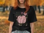 A woman with red hair wearing a black t-shirt featuring a pink pumpkin design and floral accents in an autumn setting.