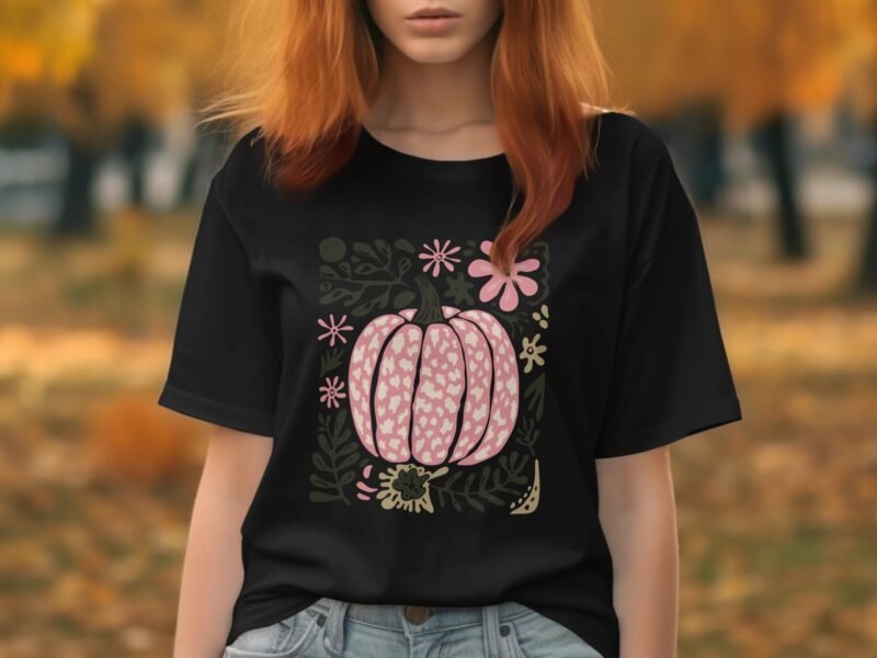 A woman with red hair wearing a black t-shirt featuring a pink pumpkin design and floral accents in an autumn setting.