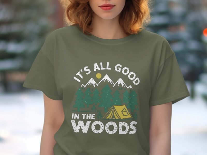Olive green t-shirt featuring "It's All Good in the Woods" graphic with mountains, trees, sun, and a tent.