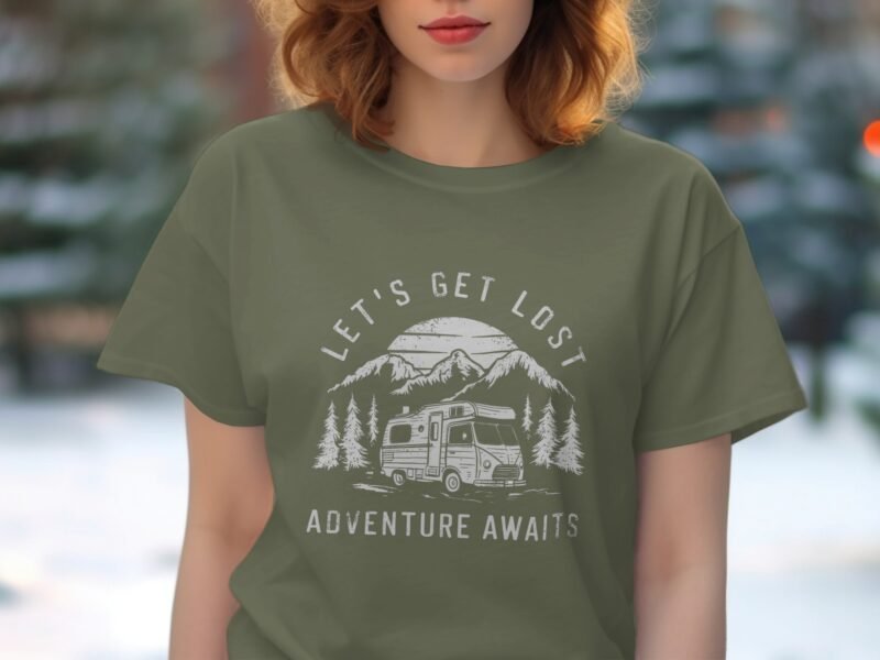 A woman wearing an olive green t-shirt with a graphic of a camper, mountains, and the text "Let's Get Lost Adventure Awaits."