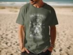 A man in a green t-shirt with a motorcycle-themed graphic stands on a sandy beach with ocean waves in the background.