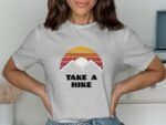 Woman wearing a gray t-shirt featuring a sunset and mountains graphic with the text "TAKE A HIKE."