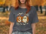 A woman in a gray t-shirt featuring orange pumpkins, a ghost, and Halloween elements stands in a fall landscape.