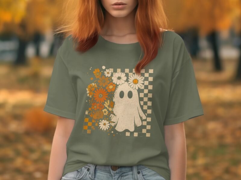 A person wearing an olive green t-shirt with a ghost and floral design, surrounded by autumn trees.