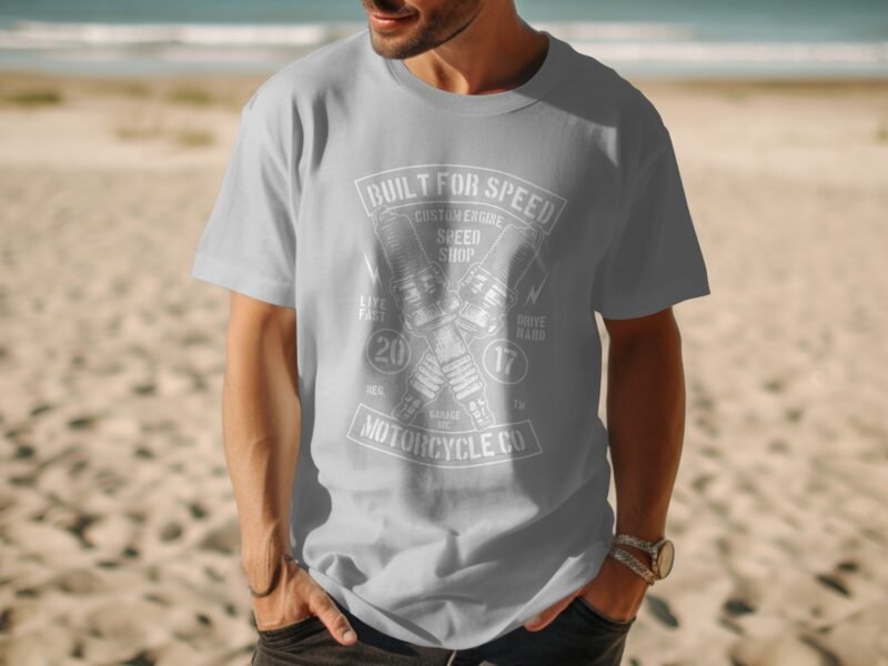 A man in a gray t-shirt with a motorcycle-themed graphic stands on a beach, with waves and sand in the background.