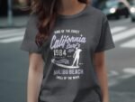 A woman wearing a gray t-shirt featuring retro surf graphics and text about California, Malibu Beach, and 1984.