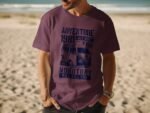 A man on a beach wears a purple t-shirt with a vintage van graphic and the text about adventure and monotony.