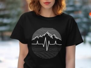 A woman with wavy hair wears a black t-shirt featuring a mountain and heartbeat graphic against a blurred outdoor background.