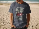 A man stands on a beach wearing a navy blue t-shirt with an aviation-themed graphic and text about flying adventures.