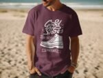 Man wearing a maroon t-shirt with a white sneaker graphic and the text "Good Shoes Take You to Good Places," beach background.
