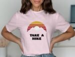 A woman models a pink t-shirt with a retro mountain and sun graphic, reading "TAKE A HIKE." Casual, cheerful style.