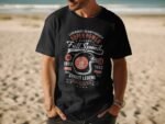 Man wearing a black t-shirt with a graphic design about motorcycle racing and street legends, standing on a beach.